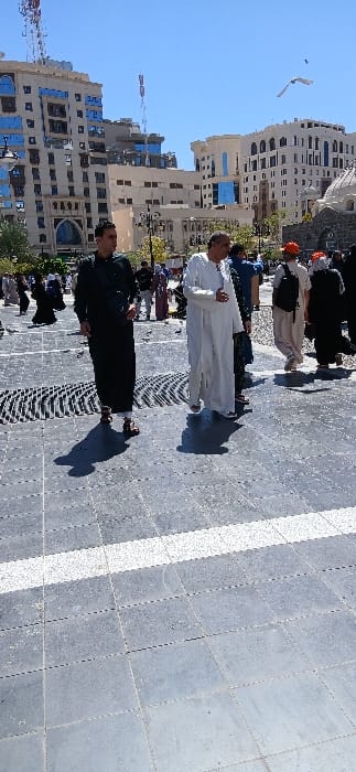 The image captures a sunny, outdoor urban scene, likely within a plaza in the city of Medina, Saudi Arabia. The foreground features dark grey square paving tiles with a distinct horizontal white stripe running across the lower mid-ground and a dark, patterned metal grate on the left.

Several individuals are present, walking across the paved area. In the midground, an adult male in a dark green long-sleeved garment and brown sandals walks towards the right, his left arm slightly bent, with a dark strap across his chest. To his right, another adult male wears a white long-sleeved thobe/dishdasha, walking generally forward with his left hand slightly raised. Both men cast pronounced shadows to their left, indicating strong sunlight. Behind them and extending into the mid-distance, other pedestrians are visible, some wearing traditional attire such as abayas and head coverings, while others wear more casual clothing, including one person in an orange cap carrying a backpack.

The background is dominated by several multi-story modern buildings constructed from light-colored materials, featuring blue reflective windows and intricate architectural designs. A prominent building on the left has an arched lower facade and an antenna with red and white segments on its roof. To the far right, a building displays a small, light-colored dome structure. The sky is clear and bright blue. A single white bird is captured in flight in the upper right portion of the sky. Street lamps are visible among the buildings.