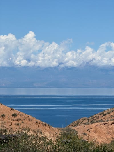 The image depicts a landscape in Tosor, Kyrgyzstan, oriented with the sky on the left and the ground on the right. On the left is a blue sky with white clouds. The center shows a wide expanse of blue water, possibly a lake, marked with distinct linear reflections. On the right, a steep, reddish-brown, earthen embankment is covered in sparse vegetation. A utility pole with wires is visible near the embankment's base, near the water's edge. The embankment continues into an area covered in denser green foliage. There are no people or animals visible in the image. The environment suggests an elevated vantage point overlooking a body of water, adjacent to a natural embankment modified by the addition of utility infrastructure.
