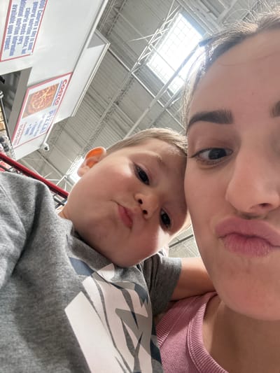A close-up, low-angle selfie shows a woman and a young child in what appears to be a large retail store, possibly Costco, given the signage in the background. The woman is on the right side of the frame, looking towards the camera with her lips pursed. The child, positioned to the left and slightly in front of the woman, is also looking at the camera with a similar puckered expression. The child is wearing a gray t-shirt with a graphic design. The background reveals overhead fluorescent lighting, a suspended ceiling structure, and promotional signage, including an advertisement for Costco pizza. The overall impression is a casual, shared moment between the two individuals.