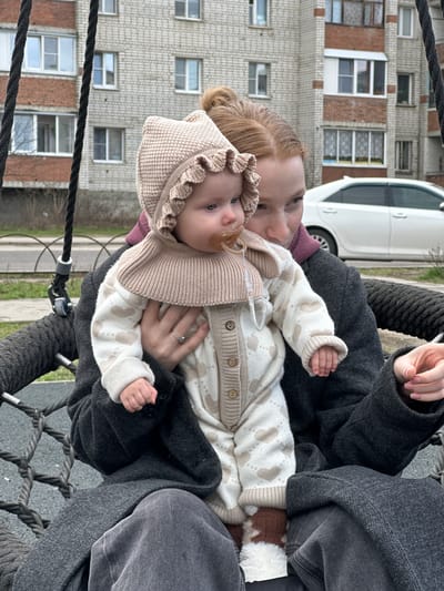 A person with red hair in a bun sits in a black rope swing holding a baby wearing a beige knit bonnet and matching cardigan and pants with a pacifier in its mouth. The baby's legs are partially covered by brown and white fur. The person wears a dark gray coat, purple undershirt, and gray pants. A building made of light gray brick and a white car are in the background. The location is Pokrov, Russia.