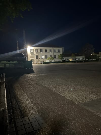 A nighttime shot depicts a building illuminated by a light, possibly a streetlight, in Nova Zagora, Bulgaria. In the foreground is a paved area, and a darker tree line or foliage is visible on the left side of the frame. The structure suggests municipal or institutional architecture. A lattice framework is visible above the streetlight, perhaps associated with it.