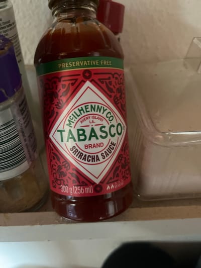 The image shows a bottle of Tabasco Sriracha sauce. The bottle is red with a green band that says "PRESERVATIVE FREE". The label has a diamond shape with "TABASCO" in large letters. There is text below it that says "BRAND SRIRACHA SAUCE". There is also text at the bottom of the bottle that says "300 g (256 ml)". To the left of the Tabasco bottle is another bottle with a purple cap and a white label with black text and a barcode. To the right of the Tabasco bottle is a clear plastic container. The items are on a shelf. There is no indication of the city of Schwäbisch Hall, Germany in the image.