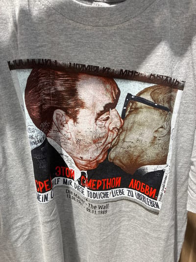 The image shows a close-up of a grey t-shirt with a graphic print. The graphic depicts a controversial 1979 painting by Dmitri Vrubel titled "My God, Help Me to Survive This Deadly Love," also known as the "Fraternal Kiss." The painting shows a socialist fraternal kiss between Leonid Brezhnev and Erich Honecker. The print on the t-shirt is detailed, showing the faces of the two men in a close embrace, with graffiti-like scribbles on their faces and surrounding areas. Below the image of the men, there is text in Russian and German. The Russian text reads "ЭТОЙ СМЕРТНОЙ ЛЮБВИ" which translates to "OF THIS DEADLY LOVE." The German text reads "HILF MIR DIESE TODLICHE LIEBE ZU UBERLEBEN" which translates to "HELP ME TO SURVIVE THIS DEADLY LOVE." Below this, there are dates "13.08.1961 - The Wall - 09.11.1989." The t-shirt itself appears to be made of cotton and is slightly creased. The background of the image is blurred, suggesting the t-shirt is laid flat or on a mannequin. No specific location in Schönefeld, Germany is identifiable from the image.