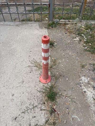 The image shows a red and white traffic cone on a patch of dirt and gravel next to a concrete sidewalk. A metal fence with circular designs is visible in the background. There is sparse vegetation and some litter on the ground. The scene appears to be an outdoor urban environment in Yangibazar, Uzbekistan.