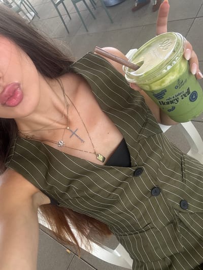 The image shows a person holding a green smoothie in a clear plastic cup. The cup has a white lid and a brown straw. The person is wearing a green and white striped jacket. There are several necklaces visible on the person's chest, including one with a cross pendant. The background suggests an outdoor setting, possibly a cafe or restaurant, with some seating visible. The lighting indicates it could be daytime. Text visible on the cup reads "Honey Veil cafe & bakery" and "Your everyday meals".