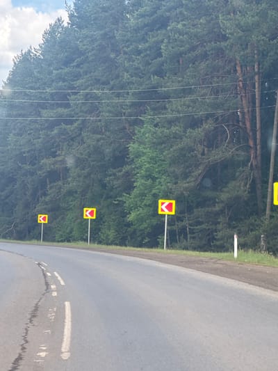 The image depicts a rural or semi-rural road scene, likely captured from a vehicle, with the road appearing to curve gently to the right as it extends into the distance. The asphalt road surface shows faint white dashed markings and a notable dark, irregular streak, possibly water or a stain, running along its length.

On the left side of the road, a narrow dirt and grass shoulder borders a dense forest of coniferous trees. Several yellow square traffic signs, each featuring a prominent red chevron pointing downwards, are mounted on white poles along the edge of the forest. At least four such signs are visible, progressively smaller in the distance, suggesting a recurring warning, possibly for a dip or hollow in the road. One additional white post, likely a roadside marker, is also visible further along the shoulder.

Several thin, horizontal wires, resembling power or communication lines, stretch across the upper portion of the tree line. A small section of blue sky with fluffy white clouds is visible in the upper left corner of the frame. The scene is illuminated by daylight, suggesting an overcast or partly cloudy sky, with no harsh shadows. No people or vehicles are visible, conveying a sense of quiet and open road. There is no discernible text in the image.