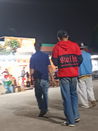 The image shows a nighttime scene with several people walking on a paved area, possibly a street or market. The lighting suggests it is dark, with artificial lights from stalls or buildings in the background illuminating the scene.
In the foreground, three individuals are walking away from the camera. The person in the center is wearing a red hoodie with black sleeves and a black rectangular patch on the back with red gothic-style lettering that reads "Star thy". Below this, there is smaller text that is difficult to decipher. This person is also wearing blue jeans and sandals. To the left of this person, another individual is wearing a blue short-sleeved shirt and blue jeans. To the right, a person in a blue jacket with yellow trim is wearing white pants and white shoes.
In the background, there are stalls or buildings with colorful signage, suggesting a marketplace or an event. Some trees are visible, and the overall atmosphere appears to be lively, despite the low light conditions. The people in the background are less distinct due to the blur and distance, but they appear to be engaging in activities at the stalls. The ground is paved, and there are some shadows cast on it, further indicating artificial lighting. The location context provided is Karangtengah, Indonesia.