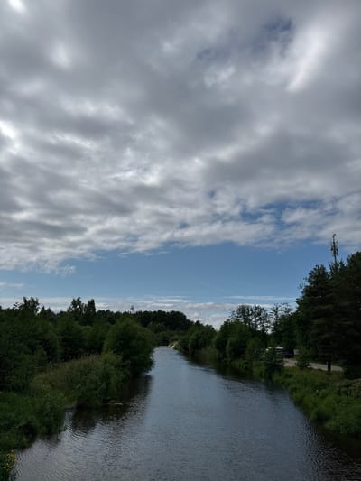 The image depicts a scenic view of the Sestroretsk River in Sestroretsk, Russia, under a partly cloudy sky. The river, with its gentle ripples, flows through a landscape dominated by lush green trees and vegetation lining its banks. On the right side of the river, a dirt road is visible, accompanied by some parked vehicles, including what appears to be a campervan and a dark car. Further along the bank, a tall telecommunications tower stands out against the sky. The sky itself is a mix of grey clouds and patches of blue, suggesting an overcast but not entirely gloomy day. The overall impression is one of a tranquil natural setting within or on the outskirts of the city.