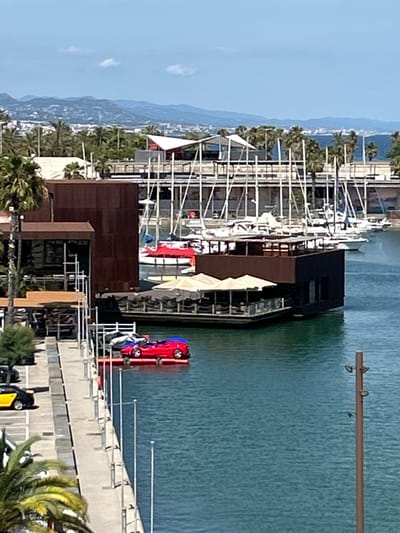 The image depicts a sunny day scene in a marina, likely in Sant Adrià de Besòs, Spain, given the distant cityscape and mountains visible. In the foreground, a concrete walkway lines the edge of a turquoise body of water. A yellow and black car is parked on the walkway, and further down, a red vehicle, possibly a car or a boat with a car-like design, is floating on the water, secured by a yellow float. Palm trees are visible along the walkway and on the land behind it.
In the middle ground, a modern building with dark brown siding and large windows is situated at the water's edge. Adjacent to it, a multi-level structure with an open-air seating area and beige umbrellas is partially submerged or floating on the water. Beyond this, a large fleet of sailboats is moored, their white sails neatly furled and masts creating a dense, vertical pattern against the sky. The background shows a hazy coastline with buildings and rolling hills under a light blue sky with scattered white clouds.