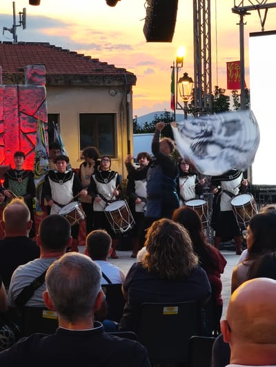 A group of young people in historical costumes are playing drums on a stage at dusk in Bojano, Italy. The sky is a mix of orange and pink hues, indicating sunset. The stage is lit with red lights on the left side. The performers are wearing vests over dark clothing and some are holding drumsticks. One performer in the center is playing a large drum, and another to the right is also playing a drum. To the right of the stage, a person is holding a large white flag with a black design. In the foreground, the backs of several audience members are visible, seated and watching the performance. A light pole with a lantern is visible on the right side of the stage.