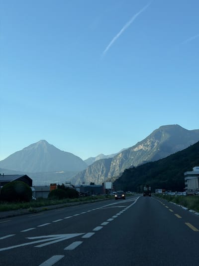 The image depicts a road leading through a valley towards mountains under a clear blue sky with a faint contrail. The road has multiple lanes with white and yellow markings. On the left side of the road, there are buildings and some vegetation, including what appear to be bushestops. Some vehicles are visible on the road, including a black car and a truck. The mountains in the background are covered in trees on their lower slopes, with rocky, barren peaks. The scene appears to be in a semi-urban or industrial area at the base of a mountainous region. The location is identified as La Balmaz, Switzerland.