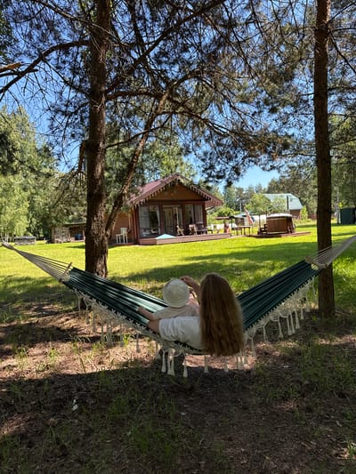 This detailed description outlines a sunny outdoor leisure scene located in Копнино, Russia.
**Scene Description:**
The image depicts a person relaxing in a dark green and white striped hammock, suspended between two prominent pine trees, observed from a low-angle rear perspective. The individual has long, light brown hair and is wearing a light-colored top and a light-colored bucket hat, with their right hand seemingly touching the hat. The hammock features white decorative tassels along its edges and is secured by rope to the tree trunks.
The immediate foreground under the hammock is shaded, showing bare earth with sparse patches of grass and fallen debris. Beyond this, a wide, brightly sunlit expanse of well-maintained green lawn dominates the mid-ground.
In the background, several wooden buildings are visible under a clear, bright blue sky. To the left, a rustic, log-style structure, possibly a wood shed, is partially obscured by trees. Centrally located is a two-story wooden house with dark brown siding, a reddish-brown gabled roof, and large windows or glass doors. A white exterior unit (likely air conditioning) is mounted on its side, and white string lights are draped along its eaves. A patio with outdoor chairs and a small table is visible in front of this house.
To the right of the central house, an outdoor dining area with a table and chairs is set up under a light-colored canopy. Adjacent to this, a wooden hot tub or outdoor bath structure is positioned on a wooden deck, with a small flag (partially obscured) flying nearby. Further in the background, another wooden house with a green gabled roof is visible, along with two dark green rectangular utility containers on the far right. The overall environment suggests a recreational or holiday property, characterized by natural elements and rural-style accommodations bathed in bright daylight, casting distinct shadows.