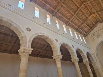 The image shows the interior of a building, likely a church or a basilica, in Abu Gosh, Israel. The architecture features a series of large stone arches supported by ornate columns. The arches are open, revealing the interior of the building with a wooden, vaulted ceiling supported by beams. Natural light streams in from tall, narrow windows positioned high on the wall above the arches. Along the wall, there are decorative circular motifs and a faded mural depicting figures, possibly angels or saints. The overall impression is one of historical and religious significance, with a focus on stone and wood construction and classical architectural elements.