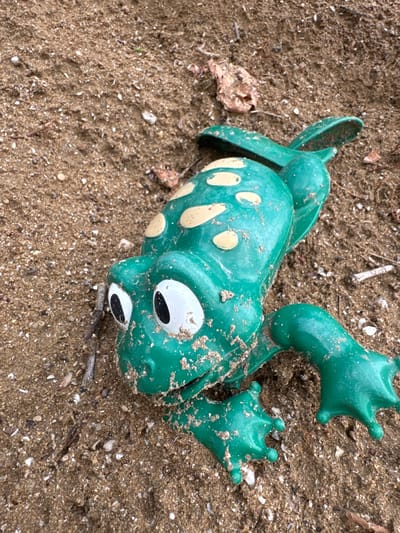 A green plastic frog toy, partially covered in sand and dirt, lies on a beach in Khazar Raion, Azerbaijan. The toy has yellow spots on its back and large white eyes with black pupils. Small shells and debris are scattered across the sandy surface. The lighting suggests an outdoor setting, possibly during the day.