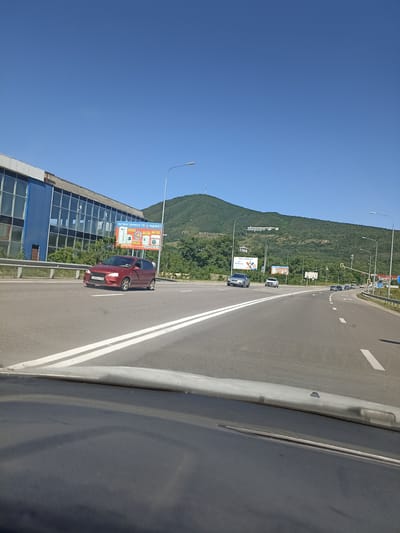 This is a daytime, outdoor, eye-level, vertical, medium shot photograph taken from a vehicle looking forward. The scene depicts a highway with clear blue skies overhead. In the foreground, the dashboard of the vehicle is visible. The highway has white lane markings and several cars are driving on it. On the left side of the road, there is a blue building with large windows. A billboard is mounted on the side of the building, displaying advertisements. Further down the road, there are more billboards and streetlights. In the background, a large, green, tree-covered mountain dominates the landscape. A communication tower is visible on the summit of the mountain. The location is identified as Goryachevodsky, Russia.