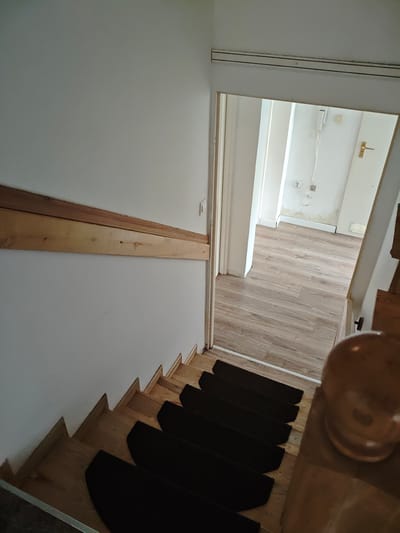 An overhead photograph captures an interior residential scene in Mława, Poland, depicting a wooden staircase descending towards an open doorway. The staircase consists of light-toned wooden steps, each fitted with a distinct, semi-circular black carpet runner. A wooden handrail, light brown in coloration, is affixed horizontally to the white-painted wall on the left side of the frame, running parallel to the descent of the stairs.
At the base of the staircase, an open doorway reveals a room or hallway beyond. This area features light-colored, wood-patterned laminate flooring. The walls in the background are painted white or off-white, displaying significant discoloration and peeling on the lower sections, particularly near the baseboards, suggesting potential water damage or neglect. Multiple electrical outlets are visible on these walls, with associated loose wiring hanging freely. A white interior door, fitted with a golden-hued handle, is positioned at the far end of this space. Near the floor on the right side of the background door, a white rectangular vent is visible. Higher on the left background wall, a white wall-mounted device, possibly an intercom or thermostat, is present with thin white cables extending from it.
Above the doorway, a horizontal track, possibly metallic or plastic, with two distinct parallel dark lines, is mounted on the wall. In the immediate foreground on the right, a portion of a rounded, wooden newel post or banister element is out of focus. No individuals or active interactions are present within the scene. The overall environment suggests a functional but somewhat unmaintained area.