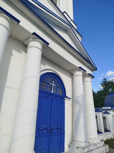 A low-angle, medium shot captures the entrance of a white building with blue accents in Мамонтово, Russia. The focal point is a double door, painted a vibrant blue, featuring an arched window above with a white cross. The building's facade is adorned with white, cylindrical columns, also capped with blue. The sky is a clear blue, sparsely dotted with clouds. In the background, a hint of greenery from trees and parts of a blue-roofed structure can be seen. A low white wall with black fencing runs along the side. The building's architecture, combined with the religious symbol on the door, suggests it may be a church or related religious building.