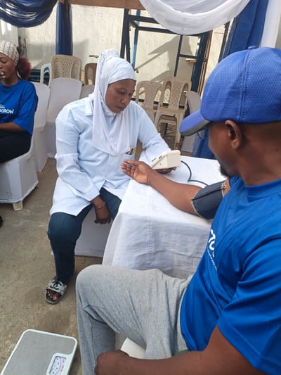 This media file depicts an outdoor community health screening event taking place during the day in Tarauni, Nigeria. The central focus is on a woman in a white lab coat and white hijab, who appears to be a medical professional, administering a blood pressure check to a man. She is seated at a white-clothed table, holding a digital blood pressure monitor, while the cuff is secured around the man's right arm. The man, seen from behind, is wearing a blue t-shirt and a blue baseball cap.

To the left of the main scene, a young woman in a blue t-shirt and a patterned headscarf is seated, looking downwards. Part of the text on her t-shirt reads "...GROW". The man's blue t-shirt also displays text, specifically "...CHILDREN SINCE 1972," suggesting an affiliation with a charitable or aid organization.

The setting is simple yet functional, featuring a temporary canopy made of white and blue fabric overhead. Several white plastic chairs, some draped in white fabric covers, are arranged around the area. A textured, light-colored wall forms the background. The ground is concrete. In the bottom left corner, a mechanical weight scale with numerical markings (0-120) is visible on the ground. The scene conveys a sense of calm and focused activity, consistent with a community health outreach effort.