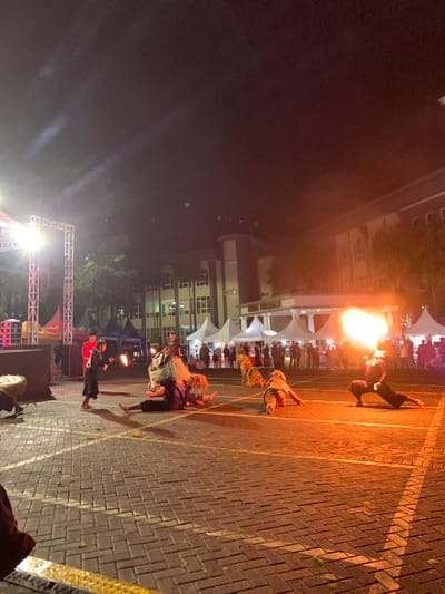 At night, a performance unfolds on a brick surface. A figure spits a burst of fire into the air, while three others crawl on the ground. Behind them, tents are set up, and people stand watching. A stage with lights and speakers is visible, suggesting a larger event. A building looms in the background.
