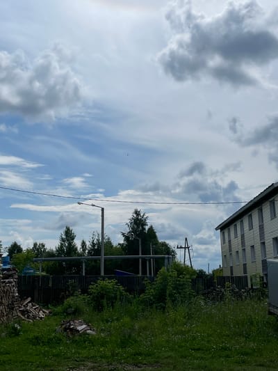The image depicts a scene in the city of Kumy, Russia, on a cloudy day. In the foreground, lush green grass with small white flowers covers the ground. To the left, a large stack of firewood is visible, along with several smaller piles of wood. A dark wooden fence runs across the middle ground, separating the grassy area from a row of trees behind it.
In the mid-ground, a tall streetlamp with two fixtures stands on a metal pole. Beyond the fence and trees, a modern, light-colored multi-story building with many windows is situated on the right side of the image. Power lines stretch across the sky, supported by a wooden pole with crossbars. The sky is filled with dramatic, grey and white clouds, with patches of blue sky visible. The overall impression is of a somewhat rural or suburban setting under a dynamic sky.