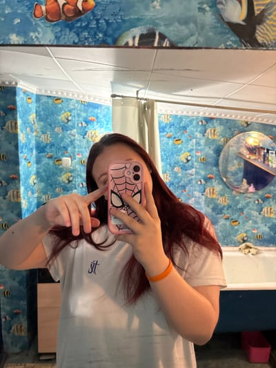 A person is captured in a mirror selfie, with the image rotated 90 degrees clockwise. The individual, who has reddish-brown hair and visible facial piercings (including a nose ring), holds a smartphone with a distinctive pink case featuring a white spider-web design and a spider emblem, resembling Spider-Man's mask. Their right hand, adorned with an orange bracelet, holds the phone pointed towards the mirror.

The person is wearing a white short-sleeved t-shirt with a small, stylized blue logo on the front, which appears to be the letters 'TJ' or similar. The background behind the person is a wall covered in an aquatic-themed wallpaper or mural, predominantly blue, depicting various cartoonish fish such as clownfish, angelfish, and other striped fish. To the right of the person's head, a round mirror is visible, reflecting a distorted view of the room, including what might be overhead lighting or another part of the wall.

To the left of the person, a white tiled or paneled wall is visible, separating the aquatic-themed area from another section of the room, which also features fish artwork. Hints from the crops show dark blue and pink elements in the further background, possibly furniture or room dividers. The setting is an indoor space, consistent with a residential or recreational area. There is no other legible text besides the t-shirt logo and the spider emblem on the phone case. The person's expression is not clearly visible, as their face is mostly obscured by the phone.