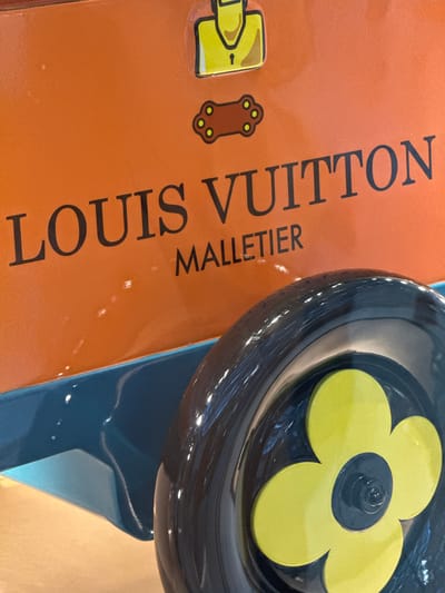 The image shows a close-up of an orange object with "LOUIS VUITTON MALLETIER" printed in black. Above the text are two cartoonish brown shapes with yellow dots, one resembling a keyhole and the other a paw print. In the foreground, a black wheel with a yellow flower design is visible, attached to a teal-colored base. The scene appears to be indoors, with reflections on the surfaces suggesting artificial lighting. The image does not provide information about a specific location in Nong Prue Subdistrict, Thailand.