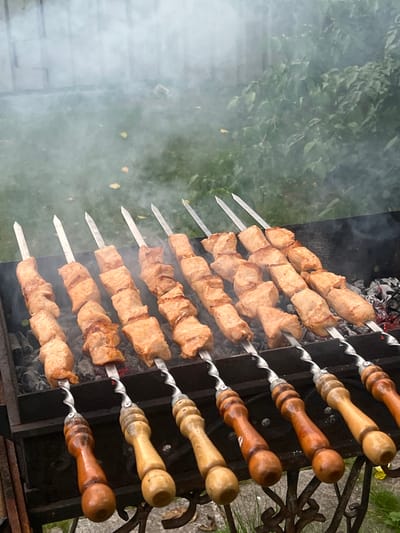 In Sukhanovo, Russia, an outdoor barbecue scene unfolds with several skewers of meat cooking over hot coals. The meat, cut into bite-sized cubes, is threaded onto metal skewers, with the handles of some skewers visible and made of wood. Smoke rises from the barbecue, obscuring the background which appears to be a grassy area with some green foliage and fallen leaves. The barbecue itself is a dark metal brazier, filled with glowing charcoal. The image captures the cooking process of shashlik, a popular grilled meat dish.