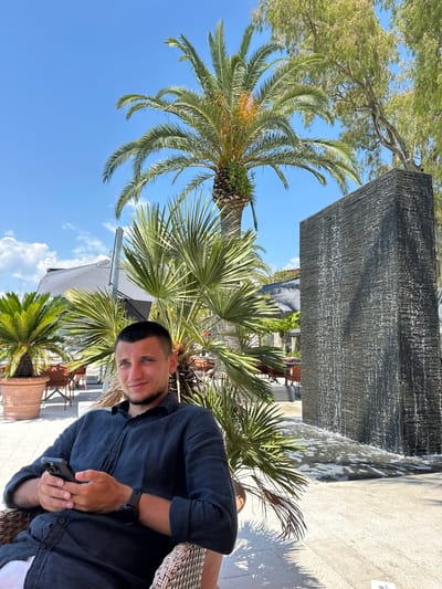 A young Caucasian man is seated in a wicker chair, holding a smartphone in his hands. He is wearing a dark, long-sleeved button-up shirt and has short dark hair and a beard. He is looking directly at the camera with a slight smile. Behind him, a large, dark rectangular structure with water cascading down its surface is visible, surrounded by lush green palm trees and other foliage. In the background, there are hints of outdoor seating and umbrellas, suggesting a resort or restaurant setting. The sky is clear blue with a few scattered white clouds. The man is in the foreground, and the water feature and palm trees are in the mid-ground and background. The overall impression is of a relaxed, sunny day in an outdoor environment. The location is specified as Donja Lastva, Montenegro.