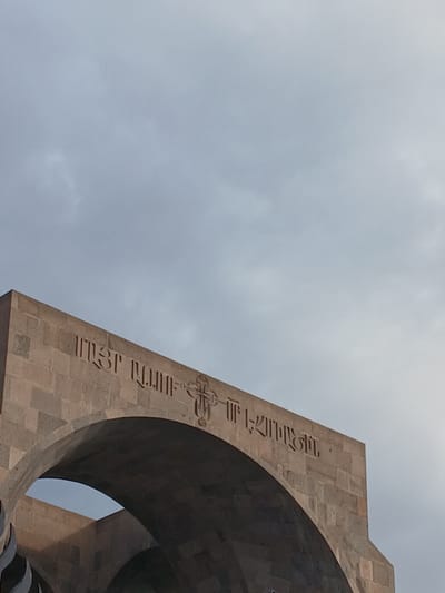 The image presents an upward-looking view of a large, light brown stone monument against an overcast sky in Vagharshapat, Armenia. The monument features a massive, curved archway or opening on its right side, which appears dark and shadowed within the lighter stone structure.

Carved into the upward-sloping face of the monument are inscriptions in Armenian script. Part of the visible text reads "ՍՈՒՐԲ ՂՈՒԿԱՍ" (Surb Ghukas), which translates to "Saint Luke." Further along the carved line, above an ornate Armenian cross symbol, the text "ԵՂԲԱՅՐԱՍԻՐՈՒԹՅՈՒՆ" (Yeghbayrasirut'yun) is visible, meaning "Brotherly Love" or "Fraternity." The intricate cross symbol is integrated directly into the text line.

The background is a uniform expanse of cloudy, light grey sky, indicating an overcast day. No direct sunlight is apparent, contributing to a somewhat solemn or diffused lighting quality. There are no people or discernible activities within the frame, placing full focus on the architectural details and the symbolic inscriptions of the monument. The choice of stone and the Armenian script and cross are distinctive cultural and geographical markers.