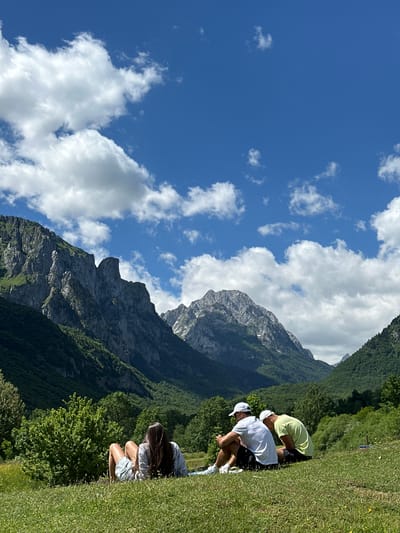 In Vusanje, Montenegro, three individuals are seated on a grassy hillside, facing away from the camera and towards a valley with lush green trees and imposing, rugged mountains. The sky is a vibrant blue, dotted with fluffy white clouds. A woman in a striped shirt and shorts reclines in the foreground, while two men in casual attire, one in a white t-shirt and baseball cap, the other in a lime green t-shirt and baseball cap, sit beside her. They appear to be relaxed, possibly engaged in conversation or enjoying the scenic vista.