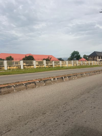 The image captures a view of a street in Kona Garu, Nigeria. The foreground features a paved road with a concrete curb, showing signs of wear and tear. Beyond the road, a yellow fence with white pillars encloses a property. This fence is topped with barbed wire. Behind the fence, a building with a red corrugated roof is visible, along with some trees and a partially constructed dark-roofed building on the right side of the frame. The sky above is overcast with grey clouds. There are no people or vehicles present in the image.