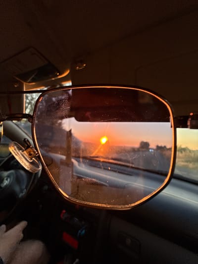 The image is an interior view from a vehicle, focusing on a rearview mirror. The mirror reflects a sunset, with the sun appearing as a bright, glowing orb in the sky. The sky in the reflection is a blend of warm oranges and soft whites, with silhouetted trees visible in the background. The mirror itself has a slightly curved, oval shape with a thin, metallic orange frame. It is attached to a mount with a suction cup, suggesting it is an accessory mirror.

The interior of the car is dark, with shadows obscuring most details. However, hints of the car's interior are visible, including part of the dashboard and a hand resting on what appears to be the steering wheel. Some dust or smudges are visible on the surface of the mirror. The overall lighting suggests it is either early morning or late evening, coinciding with the sunset reflection. The scene conveys a sense of being on a journey during a beautiful sunset. No visible text is present in the image.
