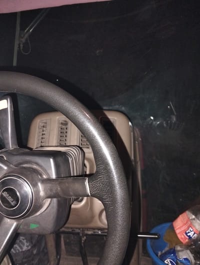 The image shows the interior of a vehicle, likely a tractor, from the perspective of the driver. A steering wheel with a "CASE II" logo is prominent in the foreground. Behind the steering wheel, a beige dashboard with vents is visible. To the right, a blue bucket contains plastic bottles, one with an orange label that reads "FANTA". The overall environment appears dark and somewhat cluttered. The scene is set within the city of Titov, Kazakhstan. There are no people visible in the image.