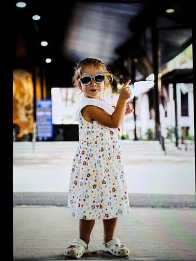 A young girl with blonde hair, wearing a white dress with colorful cartoon characters, stands on a tiled floor. She has large, novelty sunglasses shaped like shark teeth on her face and is pointing her right index finger upwards. The background is blurred, showing a covered walkway with pillars and some indistinct signage. The environment appears to be outdoors, possibly in a public space within Vientiane Capital, Laos.