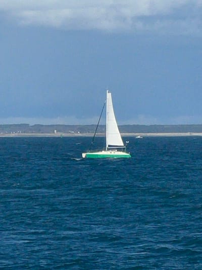 A green-hulled monohull sailboat with a white main sail unfurled is underway on deep blue, choppy water. The sailboat's hull is bright green, and its deck and cabin superstructure are white. A black mast supports the white sail. No individuals are visible on board. In the distant background, a low-lying coastline stretches horizontally across the horizon under an overcast sky, transitioning from light blue to grey. The distant landmass features a light-colored sandy or beach area, backed by a sparse line of green vegetation and scattered structures, possibly residential buildings. A slender white tower-like structure is discernible on the distant shore. A green navigation buoy is present in the mid-distance water to the left of the sailboat. A small, white, unidentified boat is visible further in the background, to the right of the main sailboat. The scene depicts maritime activity in the vicinity of Port-Louis, France.