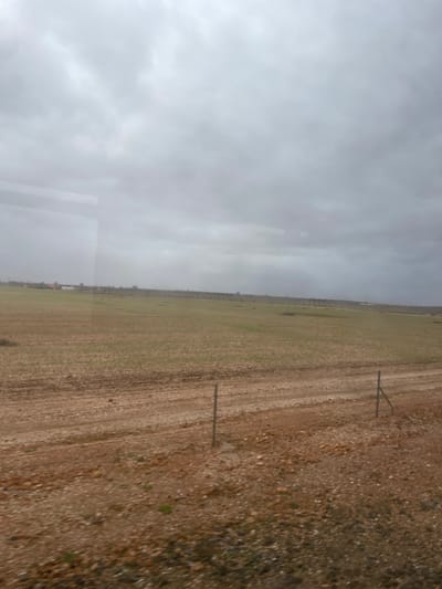 A landscape view with a cloudy sky covering much of the frame. An expansive, tilled field occupies the center, displaying rows of brown soil punctuated by sparse greenery. A narrow strip of rough, rocky terrain borders the field's edge. Sparse wooden fence posts are vertically placed along the field's border. The scene conveys a rural, possibly agricultural, setting under overcast conditions. A slight tint of a window pane is seen in the bottom left corner.