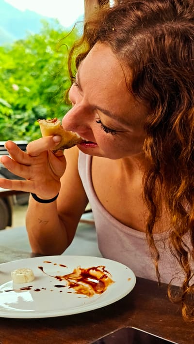 This close-up image captures a young adult woman enjoying a meal in an outdoor setting in Du Già Commune, Vietnam. The woman is shown in profile, facing left, with her upper body and head in the frame. She has auburn, curly hair and visible freckles on her face. Her eyes are slightly closed, and she is in the act of taking a bite from a food item, possibly a crepe or pastry, which she holds in her right hand. Her expression suggests enjoyment. She is wearing a light-colored tank top.

On her right forearm, a minimalist tattoo resembling the number "77" is visible, along with a dark hair tie on her wrist. A small, light-colored speck is caught in her hair near her forehead.

A white ceramic plate rests on a wooden table in the lower half of the image. The plate bears smears of a dark, glossy sauce, likely chocolate or a sweet glaze, and a small, round piece of white food, possibly a type of cheese or topping. Part of a smartphone is visible on the table at the bottom right.

The background features vibrant, lush green foliage and a bright sky, indicating a daytime scene in a natural or garden environment. The image conveys a moment of casual dining and enjoyment.
