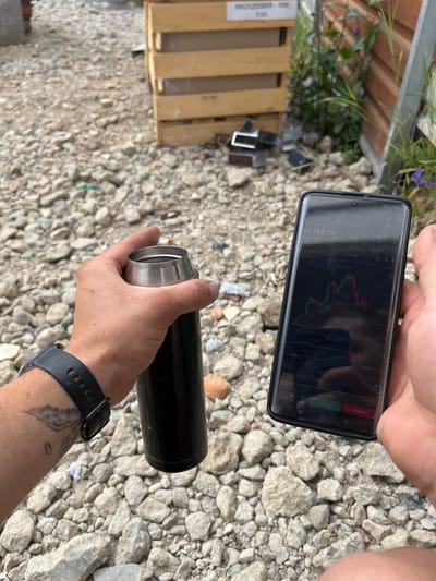 The image captures a close-up perspective of a person's left hand holding a black thermos and their right hand holding a smartphone. The smartphone displays a cryptocurrency trading interface, specifically showing a BTC/USDT chart with the current price listed as 64,118.72. The background consists of a rough, gravelly ground and a wooden structure, likely part of an outdoor setting. In the distance, a wooden crate is visible. The overall impression is that of someone checking financial data outdoors, possibly in an urban or semi-urban environment given the presence of the wooden crate and fencing in the background. There is no specific indication of the city of Jabłonna, Poland, within the image itself.