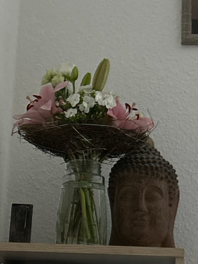 A close-up, eye-level shot shows a bouquet of pink and white lilies in a glass vase. The bouquet is nestled in a dark, woven material that resembles a bird's nest. To the right of the vase, a brown stone Buddha head is visible. The background is a textured, off-white wall. A dark, rectangular object rests on a wooden shelf to the left of the vase. The scene appears to be an indoor still life, possibly in a residential setting in Waldbröl, Germany, given the context of the request.