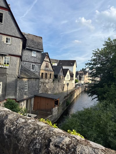 This image captures a picturesque scene of Wetzlar, Germany, likely during daylight hours, judging by the bright sky and clear shadows. The perspective is from a slightly elevated position, looking down and across a narrow waterway or canal that runs alongside a row of historic buildings.

The architecture is characterized by steep, grey slate roofs, a common feature in many European towns. The buildings are densely packed, creating a charming, old-world atmosphere. Some structures have visible stone facades, while others are painted in lighter colors, such as beige and cream. Windows, some with white frames, are scattered across the buildings, offering glimpses into the interiors or reflecting the sky. A few trees, one particularly lush and green, frame the scene and provide a natural contrast to the urban environment.

In the foreground, a rough, textured stone wall juts into the frame, suggesting the viewer is standing on a bridge or a similar elevated structure overlooking the scene. The water in the canal appears calm, with a slight ripple on its surface. A stone pathway or embankment lines the canal, with some decorative plants or flowers visible. In the distance, more buildings are visible, ascending a gentle slope under a partly cloudy blue sky.

There is no discernible activity or event taking place; the image seems to be a still shot of a tranquil moment in this historic German town. The overall mood is one of serenity and historical charm, with a clear and pleasant day enhancing the visual appeal of the location. No text is visible in the image.