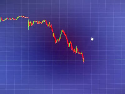 The image displays a close-up of a computer monitor showing a financial chart with a downward trend. The chart consists of red and green candlesticks against a dark blue grid background. There is no discernible location in Busmantsi, Bulgaria, as the image is of a digital display. There are no people or specific objects other than the chart itself and a mouse cursor in the shape of a pointing hand. The overall scene is an abstract representation of financial data.