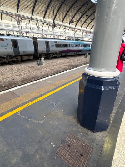 An interior view of a railway platform at Newcastle Central Station in Newcastle upon Tyne, United Kingdom, shows a stationary train and structural elements.
In the right foreground, a large, grey cylindrical column with a white decorative ring at its base transitions into a dark blue octagonal lower section. Adjacent to the column's base on the platform floor, a square, textured metal access cover with "FOR LIFTING" partially legible is present. The back of a person wearing a bright red coat is partially visible behind the column, facing away from the camera.
The platform surface is dark grey, featuring a continuous yellow safety line running parallel to the tracks. Lighter grey tactile paving sections are visible near the platform edge. Beyond the platform, two sets of railway tracks are laid on a gravel ballast bed. An electrical connection box with coiled cables stands between the platform and the tracks.
A stationary TransPennine Express Class 802 passenger train, identified by unit number 802214, occupies the tracks. The train features a silver/grey body with a distinct purple and teal/light blue stripe along its side, and "TransPennine Express" branding is visible.
The background reveals the station's high, arched, structural roof composed of metal trusses and panels, with overhead electrical catenary lines and multiple suspended light fixtures. The scene is illuminated by ambient daylight.