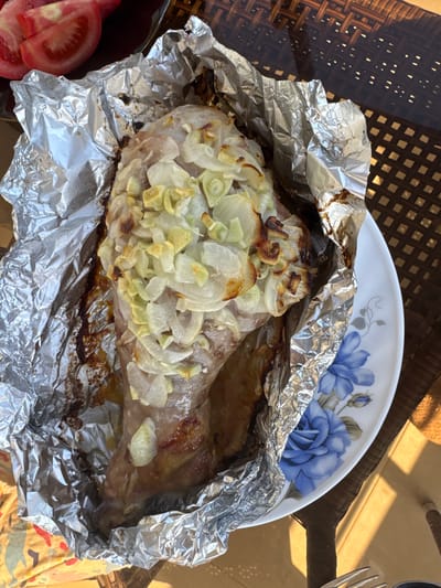 A large piece of roasted meat, likely a leg, is the central subject, served directly in its cooking foil on a white ceramic plate. The meat is generously topped with sliced onions and possibly garlic, which show softening and some browning from the roasting process. The aluminum foil is crumpled and stained with cooking juices, indicating the dish has either just been served or is being kept warm.
The white ceramic plate features a distinct blue floral pattern depicting roses and leaves. In the bottom left corner, several slices of fresh red tomato are visible, suggesting a side accompaniment. The meal is placed on a dark brown or black woven table, possibly rattan, which casts strong square-patterned shadows, indicating bright natural sunlight. A metal fork is partially visible on the right side of the frame. The strong light and woven table suggest an outdoor dining setting, consistent with locations like Erdemli, Turkey, where outdoor meals are common. The scene captures a home-cooked, substantial meal ready to be enjoyed during the daytime. No visible text is present in the image.