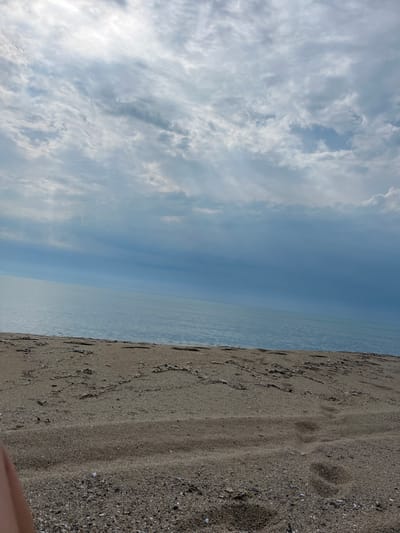 This full-frame vertical image captures a serene coastal scene in Byala, Bulgaria, featuring a sandy beach, a vast body of water, and an overcast sky. The upper half of the frame is dominated by a dense covering of grey and white clouds, with areas of brighter light suggesting diffused sun behind the cloud cover. A distinct, slightly diagonal horizon line separates the sky from the calm, light blue-grey water, which occupies the middle ground and extends horizontally across the frame. The water surface appears smooth with minimal visible texture. The bottom third of the image shows the sandy beach, characterized by light brown granular sand intermixed with darker patches of small pebbles or shells. Subtle undulating patterns and several discernible footprints are visible on the sand. A portion of a light-skinned human limb is partially visible in the bottom left corner of the frame, indicating the presence of an individual outside the main view. No other subjects, objects, or explicit actions are present in the scene.