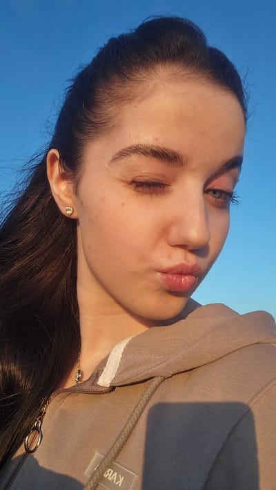 A young female subject is depicted in a close-up, head-and-shoulders portrait against a clear blue sky in Buhovtsi, Bulgaria. She is winking with her right eye (viewer's left) and pouting her lips, with her head slightly tilted downward and to the left. She has fair skin, dark eyebrows, and dark, long hair that is pulled back from her face, with some strands falling over her left shoulder. Her visible left eye (viewer's right) has a light green or hazel iris, with long dark eyelashes. A small reddish blemish is present on her forehead above the right eyebrow, and a small mole is visible on her right cheek. She is wearing a light beige or taupe hooded sweatshirt with a partially unzipped front, revealing a white lining inside the hood collar and a grey drawstring. A small, light-colored rectangular patch with mirrored text "ЯАK" (likely "KAA" or "KAB" when unmirrored) is visible on the lower left front of the sweatshirt. A small, possibly diamond or crystal, stud earring is in her right earlobe, and a thin gold-colored chain necklace with a small, dark, possibly animal-shaped pendant hangs around her neck. A gold-colored ring is attached to the zipper pull of her hoodie. Strong, warm natural light, consistent with late afternoon or early morning sun, illuminates her face and upper body from the viewer's right, creating soft shadows and a slight sheen on her forehead and nose.