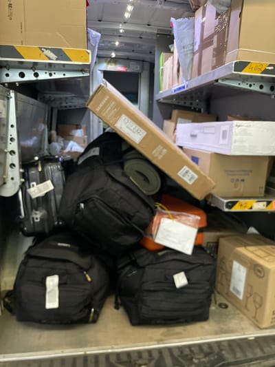 The image depicts the interior of a delivery vehicle, likely a van, filled with various packages and luggage. The items are densely packed on shelves and the floor. On the top shelf, several cardboard boxes are visible, some with labels and tape. One box in the foreground is long and narrow, with "SAMSUNG Crystal UHD" text. Below the top shelf, more boxes are stacked, including one with the number "402" on a yellow and black striped marker. In the lower portion of the image, several black backpacks are piled up. There are also some hard-shelled suitcases, one of which is dark gray or black with white labels. An orange plastic container is also visible amongst the luggage. The overall environment suggests a logistics or shipping operation. The scene appears to be from Bad Salzschlirf, Germany, though no specific landmarks are visible within the vehicle to confirm this location. There are no people visible in the image.