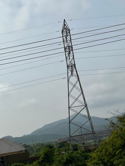 This is a medium shot, eye-level, outdoor photograph of a tall, metal electricity pylon leaning precariously to the right, set against a hazy, overcast sky. The pylon is constructed of crisscrossing metal beams and supports multiple power lines that extend horizontally across the frame. In the lower left quadrant, a portion of a building with a corrugated metal roof is visible. The foreground and midground are filled with lush green foliage, including trees and bushes. In the background, rolling, mist-shrouded hills or mountains form a distant landscape. There are no discernible people or animals in the image. The scene appears to be in a rural or semi-rural area, possibly on the outskirts of Kassa Market, Nigeria, given the presence of electrical infrastructure and the natural landscape.