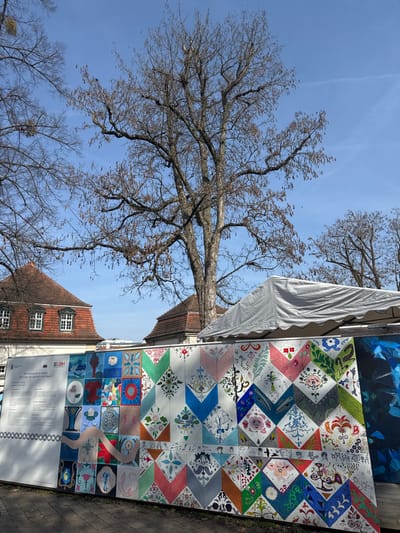 The image presents an outdoor scene in Bad Nauheim, Germany, under a clear blue sky. In the immediate foreground, a temporary, multi-panel display board is positioned. The leftmost portion of this board is a white informational panel featuring German text and multiple logos, including "LBH," with a repeating circular pattern along its lower edge. The central and right sections of the board are covered by a mosaic of numerous square panels, individually decorated with diverse, colorful designs, predominantly floral, abstract, and geometric. Many panels are arranged to form larger chevron patterns in various hues. A distinctive wavy, light-colored element traverses some of the central panels. To the far right, a large, abstract artwork, primarily blue and black, is partially visible.
In the midground, a white market-style tent with a grey tarp roof stands behind the right side of the display board. Behind the display, two buildings with traditional German architecture are partially visible. The building on the left features a red-tiled gable roof with multiple white-framed, multi-pane windows. A similar red-tiled roof is visible from the building to its right.
The background consists of a clear, bright blue sky containing a faint white aircraft contrail in the upper right. Several large, bare deciduous trees are prominent, indicating a non-foliage season. The largest tree, centrally located, exhibits mottled grey bark and numerous hanging seed pods or catkins from its branches. A tree on the far left shows visible mistletoe clusters. No people are present in the observed scene.