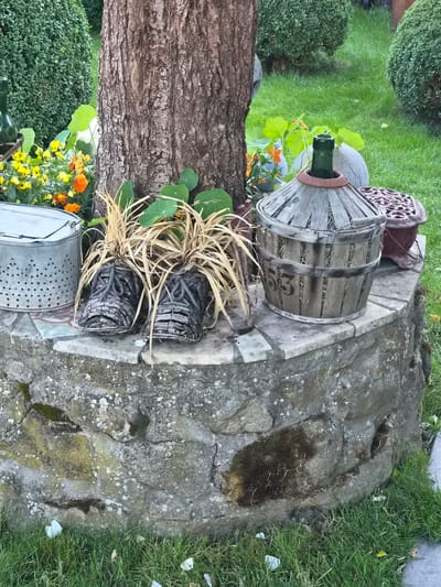 The image depicts a close-up of a garden display situated near the base of a mature tree. The display is located in the municipality of Bettembourg, Luxembourg.
The scene features a stone-constructed circular structure that forms a raised platform. On this platform, several decorative objects are arranged. To the left, a cylindrical metal container with perforations is visible. Adjacent to it are two planters shaped like shoes, constructed from a woven material and containing dried grass-like plants. Further to the right on the platform is a large, wooden, barrel-like object with metal bands, and the number "53" is stenciled on its side. Behind this barrel, a dark red, ornate metal object, possibly a decorative piece or a small grill, is partially visible. A green glass bottle is placed within the wooden barrel structure.
The ground surrounding the stone platform is covered in green grass. In the background, several meticulously trimmed green bushes are visible, suggesting a well-maintained garden. The tree trunk, with its textured bark, is a prominent feature in the center of the image, providing a backdrop to the display. The overall atmosphere is one of a garden or outdoor decoration, likely in a residential or public park setting in Bettembourg. There are no people or animals present in the image.