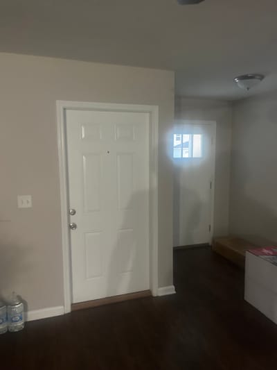 The image depicts an interior scene, likely a hallway or entryway, within a residential property. The dominant elements are two white doors, one positioned centrally and the other further down a corridor to the right. The central door is a solid panel door with a peephole and a silver doorknob and deadbolt. To the left of this door is a light switch plate on a plain, light-colored wall.

The door on the right is partially obscured by a bright glare from an unseen light source, but it appears to be a single-panel door with a glass insert at the top featuring a grid pattern. Outside this door, parts of houses can be seen through the glass, suggesting it leads to the exterior or a well-lit area.

The floor is dark, hardwood or laminate. In the foreground on the left, two plastic water bottles are visible near the base of the wall. In the background on the right, there is a long, flat object wrapped in cardboard or brown paper, and a white box with red detailing. A ceiling light fixture is visible in the upper right portion of the image. The walls are painted a neutral, light color. The overall impression is of a clean, unadorned interior space. No specific location in Jacksonville, United States of America can be determined from the visual information provided.